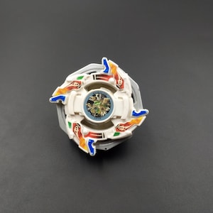 Puede incluir: Un juguete de peonza de plástico blanco, azul, rojo y naranja con un centro transparente y un anillo de metal plateado alrededor del centro. El juguete tiene cuatro aspas con un diseño de llamas.