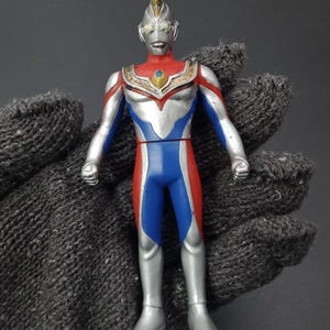 May include: A vintage action figure of a superhero with a silver helmet and body, red and blue accents, and a gold chest emblem. The figure is posed with arms at its sides, showcasing its classic design and vibrant colours.