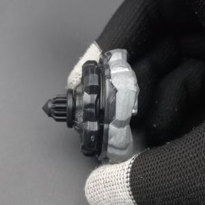 Pu&ograve; includere: Una trottola giocattolo nera e grigia con un design strutturato. La parte superiore ha una punta e una struttura a forma di ingranaggio. Il giocattolo &egrave; tenuto da una mano guantata su uno sfondo grigio.