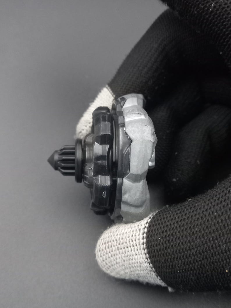 Pu&ograve; includere: Una trottola giocattolo nera e grigia con un design strutturato. La parte superiore ha una punta e una struttura a forma di ingranaggio. Il giocattolo &egrave; tenuto da una mano guantata su uno sfondo grigio.