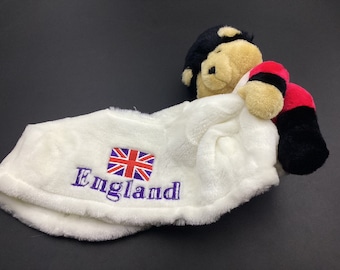 May include: White plush toy with a black and red jersey and a black head. The toy is lying on a white plush blanket with the word "England" and a red, white, and blue flag embroidered on it.