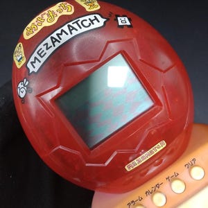 以下が含まれることがあります： 長方形の画面を持つ赤いMEZAMATCH携帯型電子ゲーム。ゲームには一連のボタンと日本語のテキストがあります。ゲームの上部には、漫画のステッカーが付いた白いバナーに「MEZAMATCH」というテキストがあります。