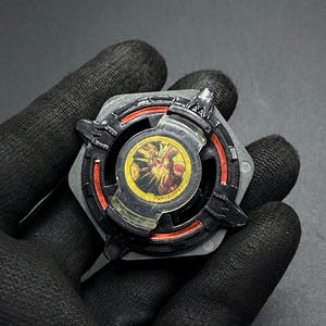 K&ouml;nnte beinhalten: Schwarzer und roter Beyblade mit einem durchsichtigen Plastikkern, der ein gelbes und rotes Design zeigt. Der Beyblade hat eine sechseckige Form mit einer spitzen Spitze.