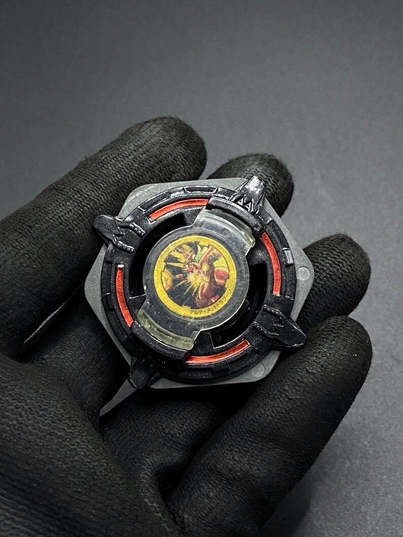 K&ouml;nnte beinhalten: Schwarzer und roter Beyblade mit einem durchsichtigen Plastikkern, der ein gelbes und rotes Design zeigt. Der Beyblade hat eine sechseckige Form mit einer spitzen Spitze.