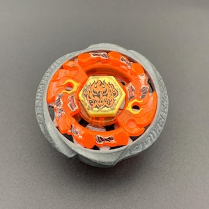 Puede incluir: Un juguete de peonza circular con un anillo exterior metálico gris y una pieza central de plástico naranja brillante. El perno hexagonal amarillo central muestra un gráfico de llama estilizado y el texto PHOENIX en negro.