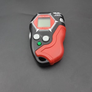 May include: A black and red handheld device with a small screen and buttons. The device has the words "DIGITAL CODE" and "SCAN-SYSTEM" printed on it. The device is likely a vintage electronic toy or game.