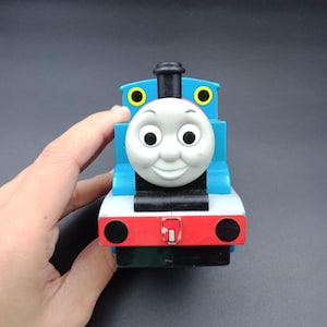 Puede incluir: Un tren de juguete azul, rojo y blanco con una cara sonriente. El tren de juguete tiene círculos amarillos en los lados y una chimenea negra. El tren es sostenido por una mano.