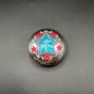 Puede incluir: Un compacto plateado con una estrella azul en el centro, rodeada de estrellas rojas. El compacto tiene una superficie reflectante y un diseño decorativo en los bordes. El artículo está sobre una superficie gris oscuro.