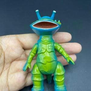 May include: A green and blue toy figure of a creature with a large open mouth, antennae, and segmented body. The figure is held in a hand against a dark background. The toy is a vibrant mix of green and blue hues.
