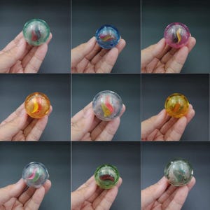 May include: Nine transparent marbles in various colours, each with a colourful, swirled design inside. The marbles are held in a hand against a dark background. Colours include green, blue, pink, orange, and yellow.