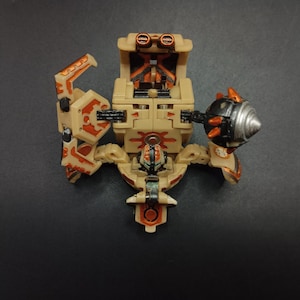 May include: Tan and orange robot toy with articulated arms and a weapon. The toy has a detailed design with visible mechanical components. The robot is positioned on a dark surface.