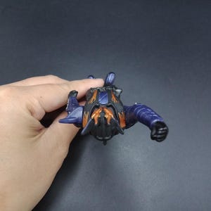 May include: A small, detailed action figure in shades of purple, black, and orange. The figure has a stylized insect-like design with articulated limbs and a textured surface. The figure is held against a dark background.