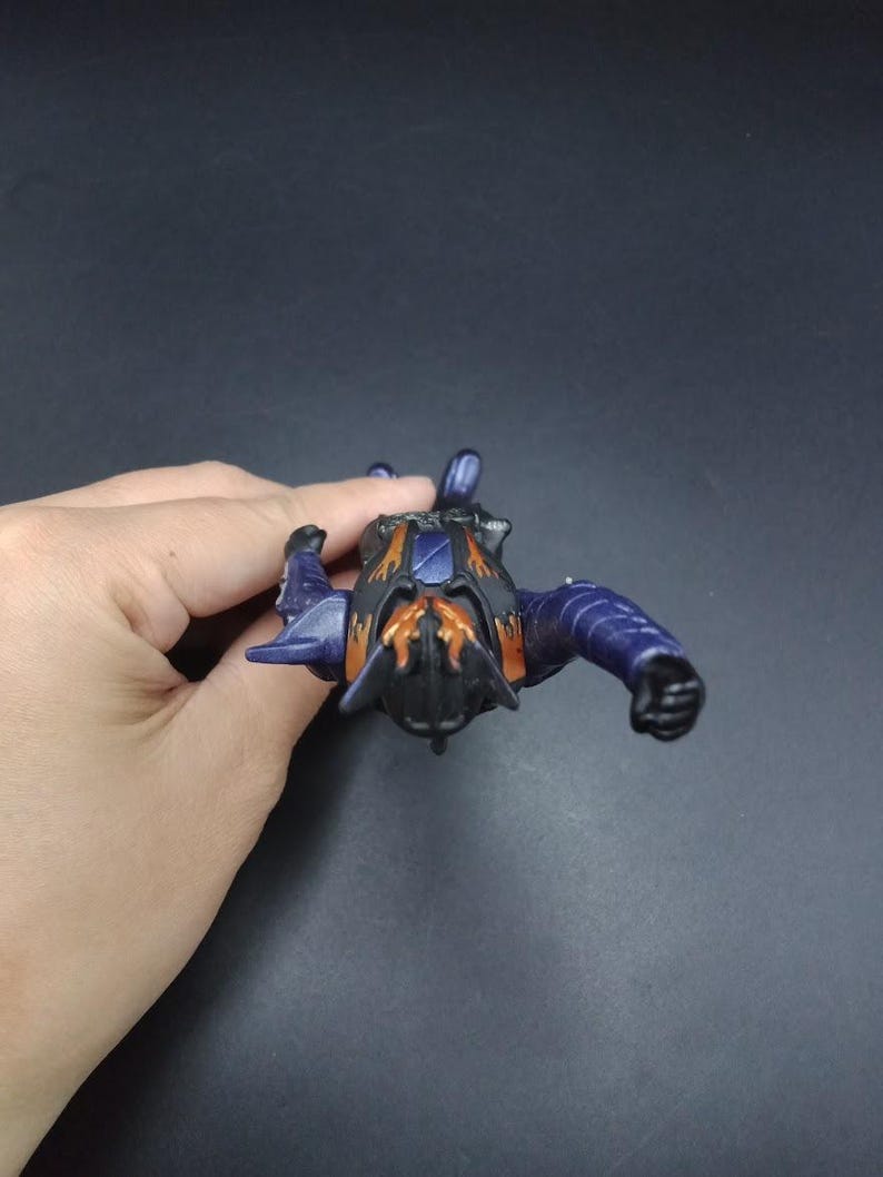 May include: A small, detailed action figure in shades of purple, black, and orange. The figure has a stylized insect-like design with articulated limbs and a textured surface. The figure is held against a dark background.