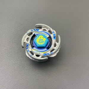 Pode incluir: Um brinquedo de pião circular com um anel exterior de metal prateado com lâminas curvas. O centro inclui um parafuso hexagonal azul com um gráfico amarelo de uma criatura estilizada e o texto "BEY" impresso numa fonte escura e negrito.