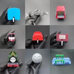 May include: A selection of toy train carriages and accessories in various colours. Includes a blue train car, a grey flatbed, a red passenger carriage, a red cargo carriage, a maroon boxcar with a face, a green engine with a face, a grey ball with a face, and a blue platform.