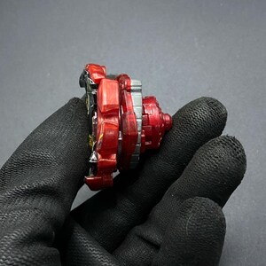 K&ouml;nnte beinhalten: Rotes, schwarzes und silbernes Beyblade mit einem roten und silbernen Kreisel. Das Beyblade besteht aus Kunststoff und Metall.