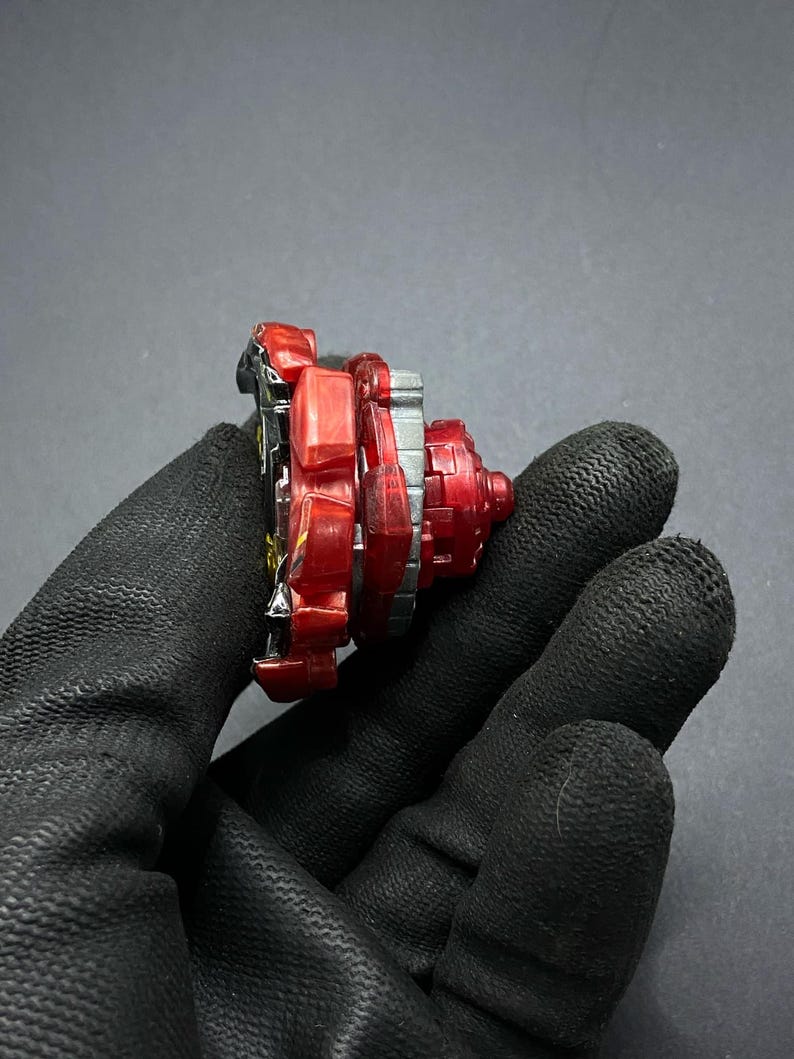 K&ouml;nnte beinhalten: Rotes, schwarzes und silbernes Beyblade mit einem roten und silbernen Kreisel. Das Beyblade besteht aus Kunststoff und Metall.