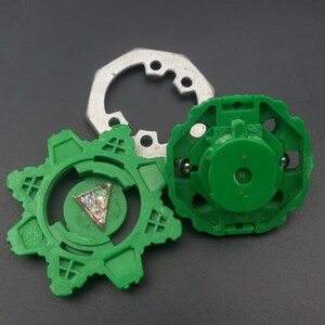 May include: Three-piece toy set featuring a green gear-shaped component, a green circular piece, and a silver metal ring. The green pieces have intricate designs and the set is on a dark background.