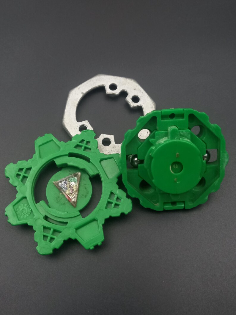 May include: Three-piece toy set featuring a green gear-shaped component, a green circular piece, and a silver metal ring. The green pieces have intricate designs and the set is on a dark background.
