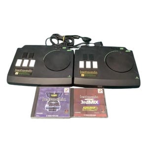 May include: Two black Beatmania DJ controllers for PlayStation, each featuring a circular turntable and three white buttons. Included are two game cases: one labeled Beatmania and the other labeled Beatmania Append 3rdMIX, with a black cable.