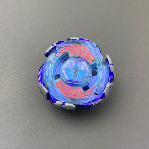 Puede incluir: Un juguete de peonza circular con un cuerpo de plástico azul translúcido y bordes exteriores de color plata metálica. El centro incluye un emblema hexagonal azul con un gráfico negro, rodeado de complejos patrones lineales rojos.
