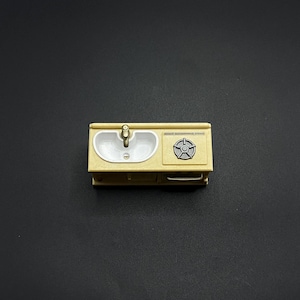 May include: A miniature beige kitchen sink and stove combination. The sink has a white basin and a gold faucet. The stove has a circular design. The miniature kitchen set is on a dark background.