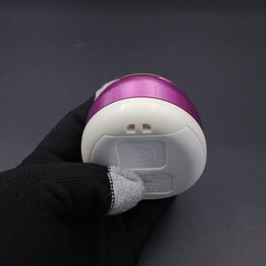 Puede incluir: Un masajeador facial el&eacute;ctrico blanco y morado. El dispositivo tiene forma redondeada con una base blanca y una parte superior morada. La parte inferior del dispositivo tiene dos peque&ntilde;os orificios y un logotipo.