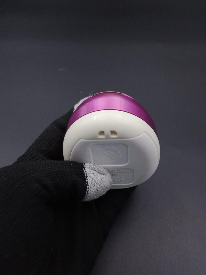 Puede incluir: Un masajeador facial el&eacute;ctrico blanco y morado. El dispositivo tiene forma redondeada con una base blanca y una parte superior morada. La parte inferior del dispositivo tiene dos peque&ntilde;os orificios y un logotipo.