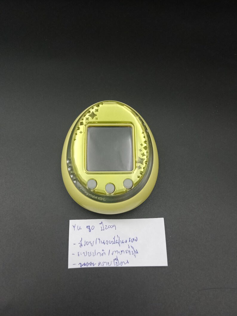 May include: A yellow handheld electronic game with a black screen and three gray buttons. The device has a gold-colored border with star-shaped designs. A handwritten note is below the device.