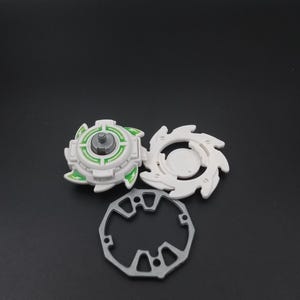 May include: A three-part toy set comprising a white and green top, a white ring, and a silver-coloured ring. The top has a circular design with green accents and text. The other white piece has a star-like design.