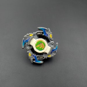 Könnte beinhalten: Ein graues und blaues Beyblade mit einem Wolf-Design auf der Vorderseite. Die Vorderseite ist weiß mit grünem Hintergrund und einem goldenen Wolf-Bild. Der Text "WOLF" ist auf der Vorderseite aufgedruckt.