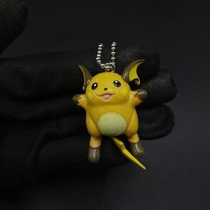 Puede incluir: Figura amarilla de Raichu con expresión alegre, pies y orejas marrones, y cola en forma de rayo. La figura está unida a una cadena de bolas plateada, posiblemente un llavero o un amuleto.