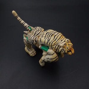 以下が含まれることがあります： うずくまる姿勢の、おもちゃのトラの置物。トラはクリーム色の体に黒い縞模様と緑色のアクセントがあります。頭にはオレンジと黒の模様が施されています。尾は伸びており、置物は暗い表面にあります。