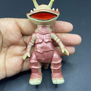 May include: A vintage toy figure of a creature with a large open mouth, two yellow-tipped antennae, and a pinkish-red body. The figure has a detailed design, with visible textures and a retro aesthetic, held in a hand.