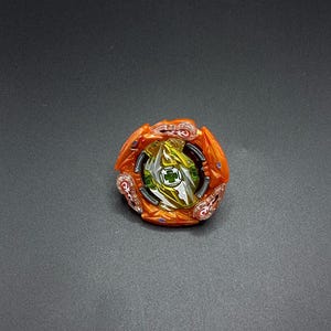 May include: A colorful spinning top toy with orange, yellow, green, and silver accents. The toy has a multi-layered design with a central green emblem and is set against a dark background.