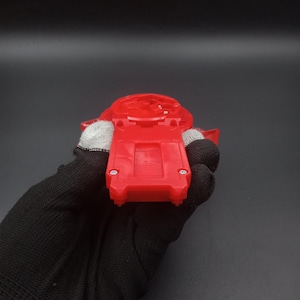 May include: A red plastic toy component, held in a gloved hand, against a dark background. The toy has a rectangular base with rounded edges and a circular top. The component appears to be a part of a larger toy.