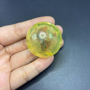 May include: A transparent yellow sphere with glitter suspended within. The sphere has three small holes and is held in a hand. The object is likely a toy or decorative item.