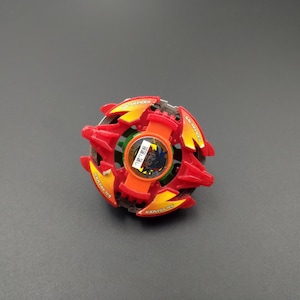 May include: A red and yellow toy with a circular design. The toy has a central orange piece with Japanese text and a black and blue graphic. The outer edges are red with yellow accents. The toy is on a grey surface.