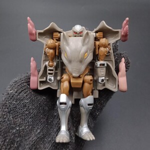 May include: A brown and gray action figure in a rat-like form, with silver legs and pink accents. The toy has a detailed design with visible joints and a small head with red eyes. The figure is a collectible toy.