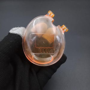 May include: A clear, peach-colored, egg-shaped container with two orange protrusions and an orange rim. The container has a rectangular section with three vertical lines. The container is held by a gloved hand.