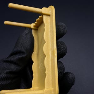 May include: A small, mustard-colored decorative shelf with a scalloped edge and two horizontal dowels. The shelf is held by a gloved hand against a dark background. The shelf appears to be made of plastic or a similar material.