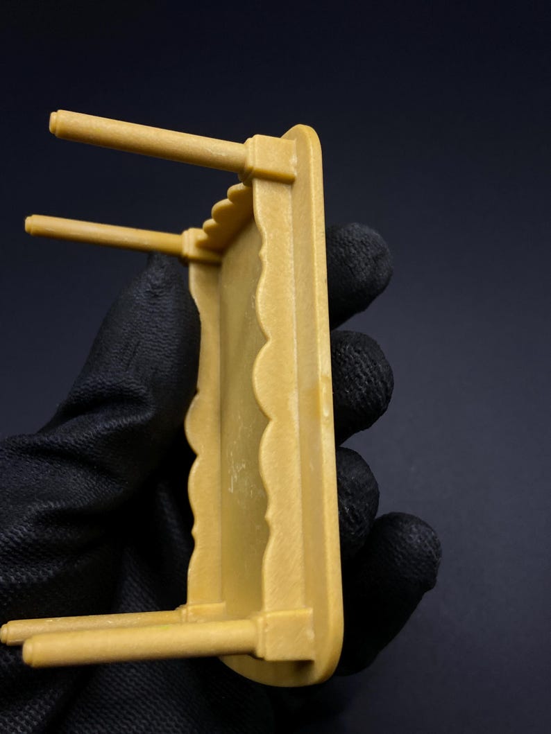 May include: A small, mustard-colored decorative shelf with a scalloped edge and two horizontal dowels. The shelf is held by a gloved hand against a dark background. The shelf appears to be made of plastic or a similar material.