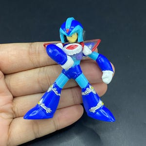 May include: A blue, white, and red action figure of a character with a helmet and armor. The figure has a dynamic pose with arms bent and legs spread. The figure is held in a hand against a dark background.
