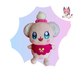 Puede incluir: Un juguete de peluche rosa con una cabeza de glaseado blanco y rosa, ojos azules y un babero rosa con un diseño de cuchara y tenedor amarillo. El juguete sonríe y tiene una cola blanca y esponjosa.