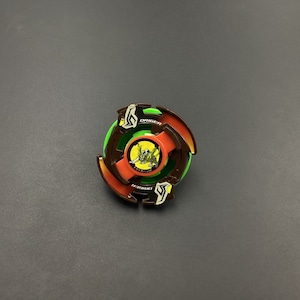 May include: A brown and green Beyblade with a yellow and black design. The Beyblade has a red ring around the centre and the text "Dragoon" on the side.