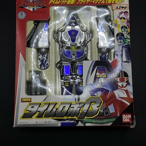 May include: A silver and blue toy robot with a red and white figure in the background. The box has the text "Time Robo B" and "Bandai 2000" on it.