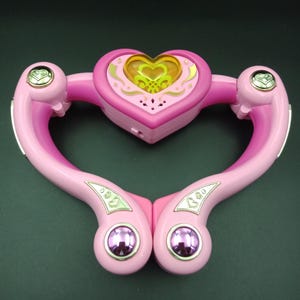 Könnte beinhalten: Ein rosa herzförmiges Spielzeug mit einem goldfarbenen Herzdesign in der Mitte. Das Spielzeug hat zwei gebogene Arme mit dekorativen Akzenten und lila kreisförmigen Knöpfen. Das Spielzeug besteht aus Kunststoff und hat eine glänzende Oberfläche.