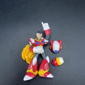 Puede incluir: Una figura de acción roja, blanca, amarilla y gris de un personaje con un mechón de pelo amarillo puntiagudo. La figura está en una pose dinámica con un brazo levantado. Se colocan piezas adicionales, incluido un casco y un componente pequeño, cerca.