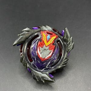 Peut inclure: Un toupie de jeu noire, rouge et violette avec un design détaillé. Le centre présente un emblème rouge et jaune avec un accent bleu. L'anneau extérieur a des lames gris foncé et des accents violets.