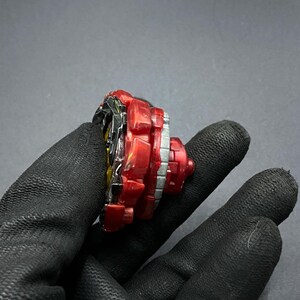 K&ouml;nnte beinhalten: Ein rotes und schwarzes Beyblade mit einer silbernen und wei&szlig;en Schicht. Das Beyblade wird in einer behandschuhten Hand gehalten.